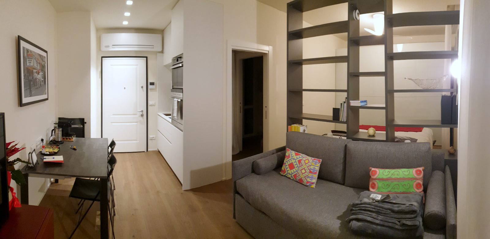 Panoramic view of the studio: dining table on the left, entrance and kitchenette in the background, sofa bed and the bookcase that screens the sleeping area on the right